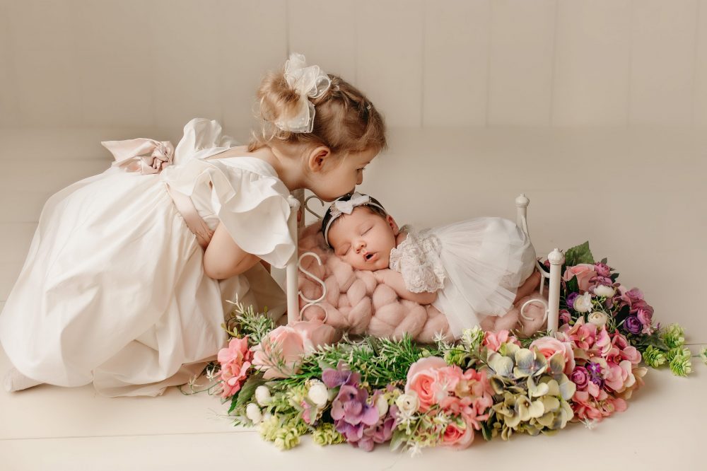 Newborn baby lying in a bed adorned with flowers, with the older sister lovingly watching over, captured during a photoshoot in Preston, Lancashire